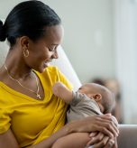 A new Mother sits in her baby's nursery with the infant in her arms and held to her chest, as she nurses him.  She is dressed casually in a yellow t-shirt and looking down at the baby with a smile.